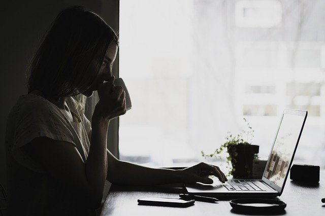 Žena sedí pri stole, pracuje na počítači a pije kávu v prítmí