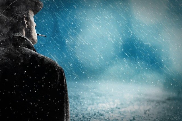Muž v klobúku a kabáte s cigaretou v ústach stojí v daždi.jpg